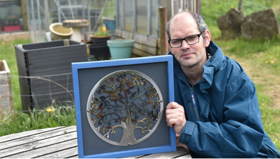 man holding a plaque he has carved out of wood sat in a garden